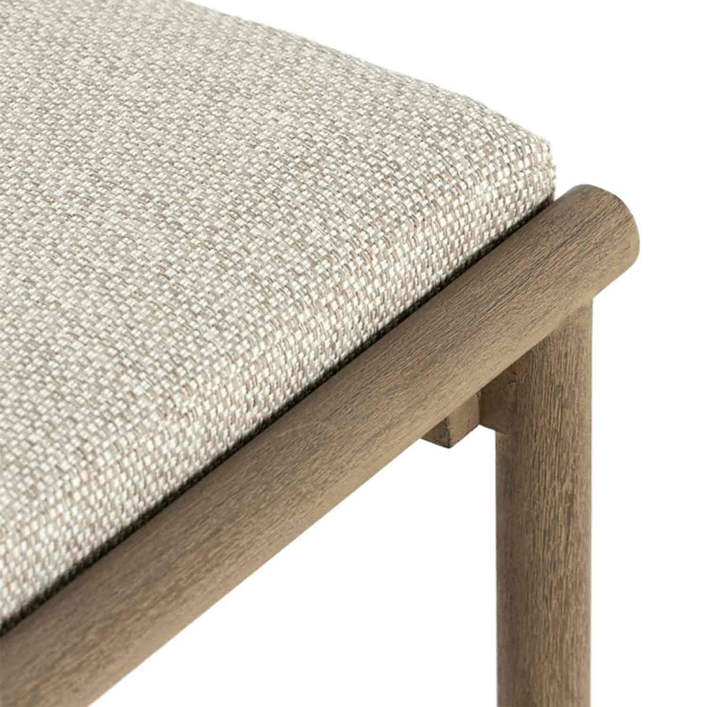 Close-up of a beige woven upholstered seat cushion on a teak outdoor bar stool, showing the textured fabric, rounded teak rail and part of the teak leg.