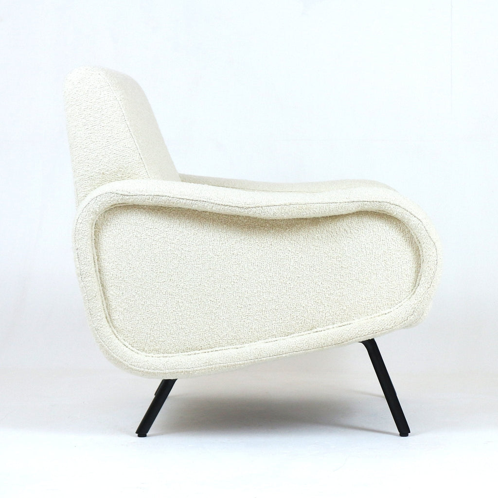 Side profile of a modern white boucle upholstered armchair with a curved low back, rounded padded arms and slender black metal legs against a plain white studio background.