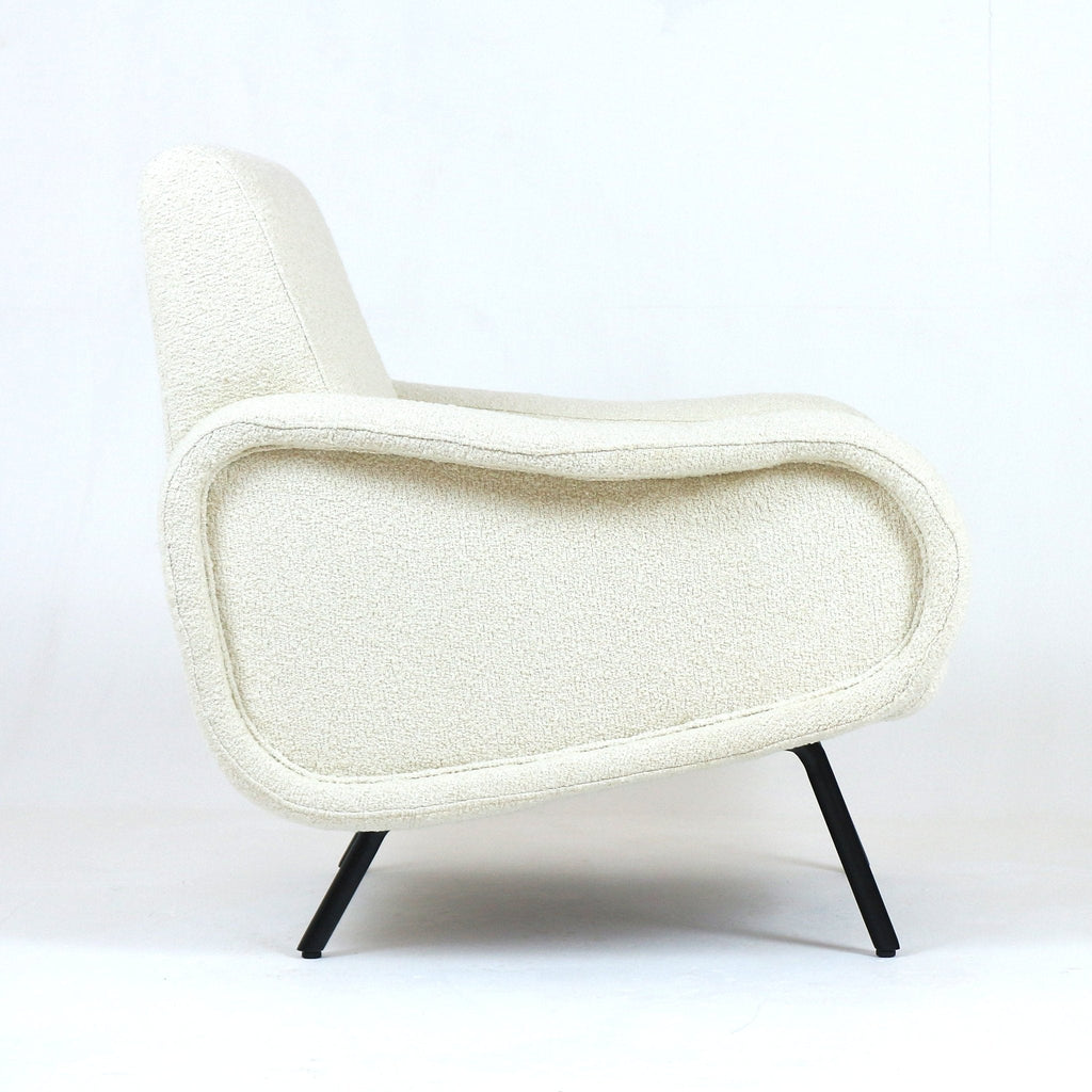 Side profile of a modern white boucle upholstered armchair with a curved low back, rounded padded arms and slender black metal legs against a plain white studio background.