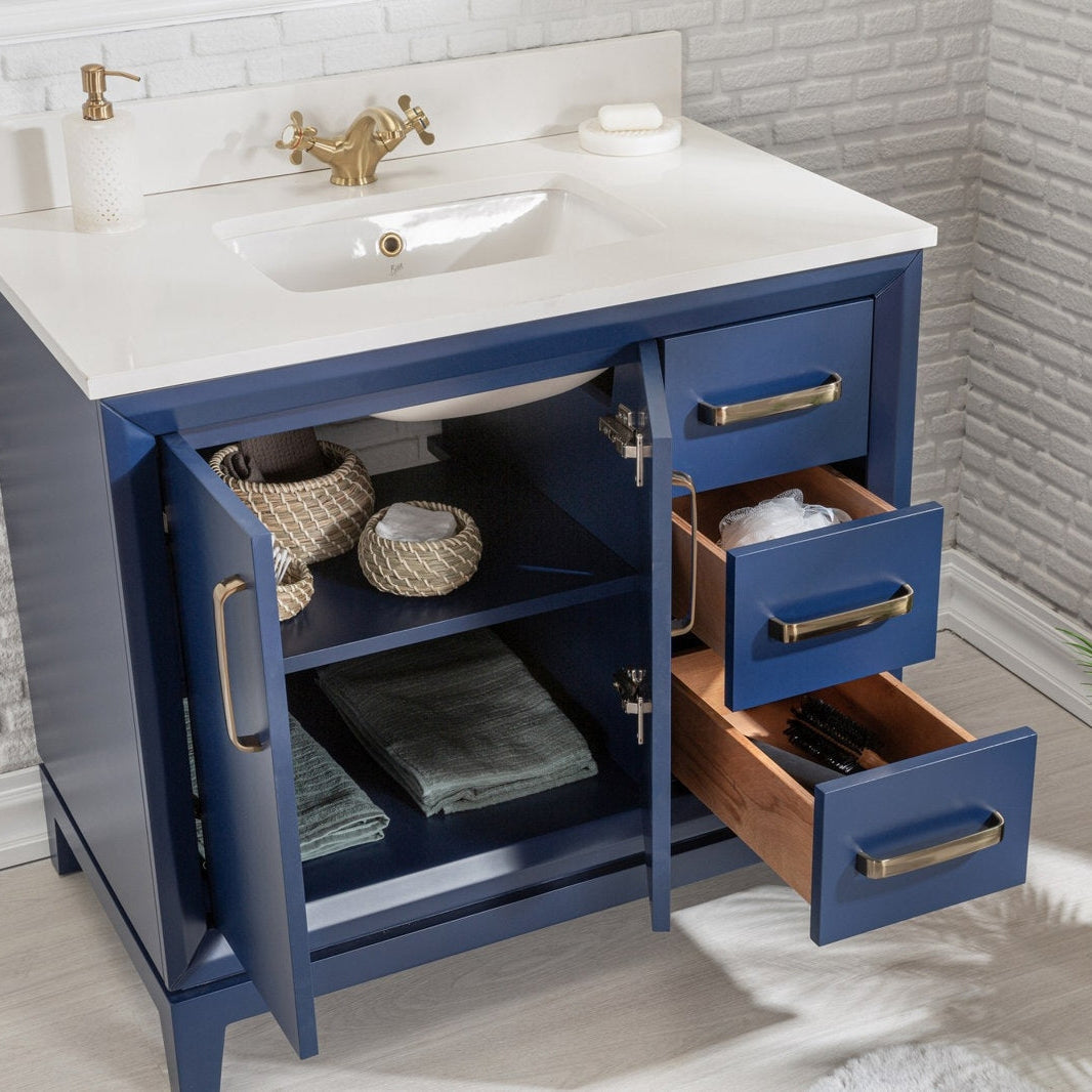 Modern navy-blue solid wood bathroom vanity with a white stone top and undermount ceramic sink, brass faucet and hardware; open cabinet and three drawers reveal woven baskets, folded towels and wood interiors.