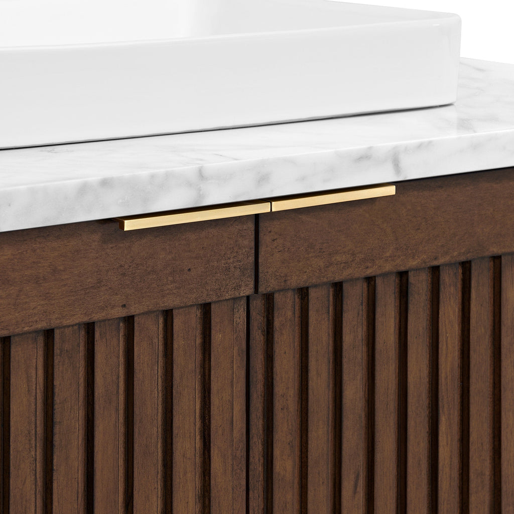 Close-up of a modern fluted solid wood bathroom vanity in warm brown, featuring a stone marble countertop, the edge of a white rectangular ceramic vessel sink, and slim brushed brass bar handles.