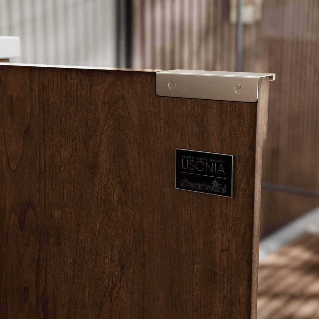 Close-up of a solid wood vanity panel in warm brown with visible grain, showing a stone top edge, brushed metal corner bracket and a small rectangular metal logo plaque mounted on the panel.