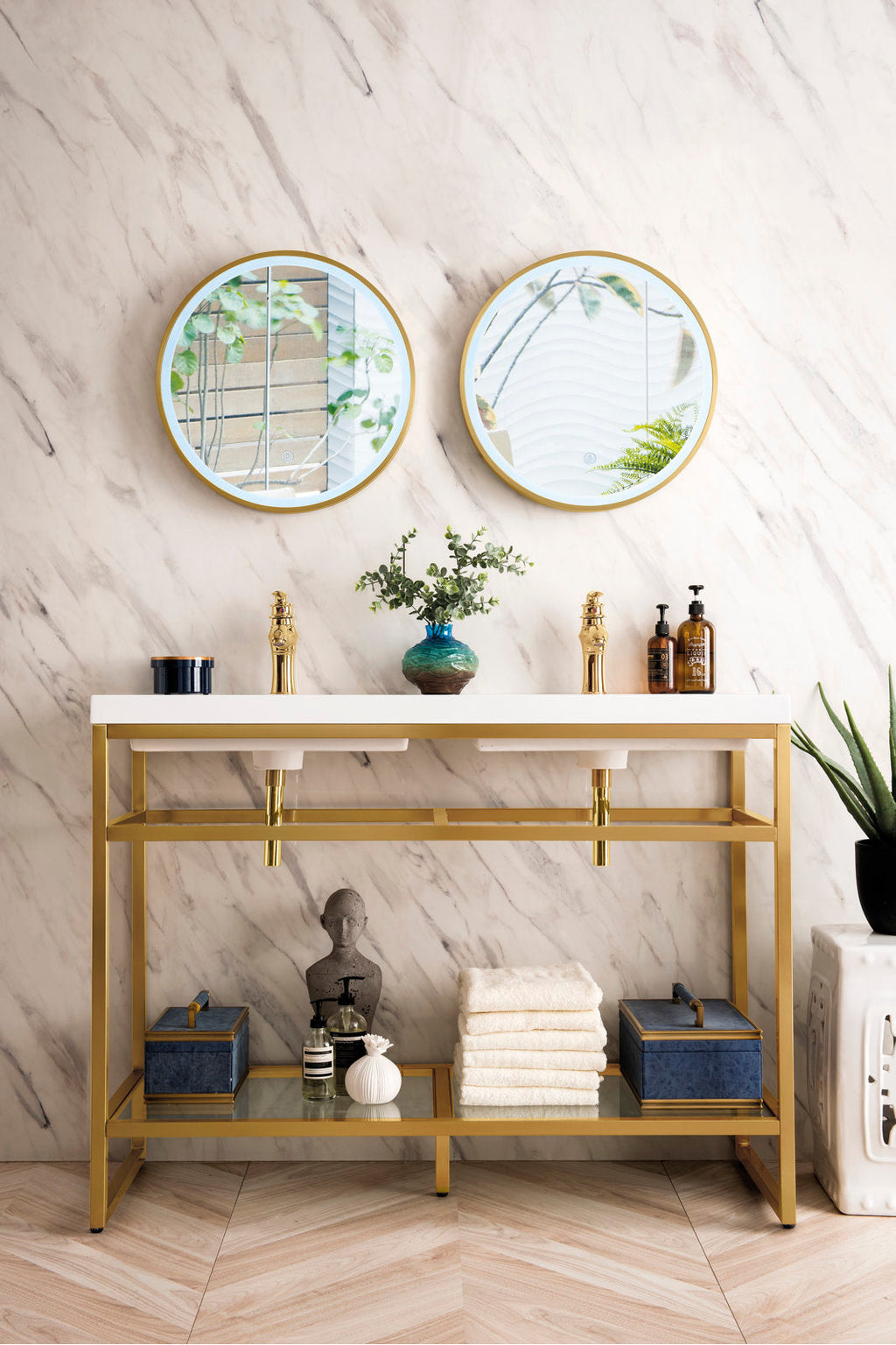 Freestanding glam-style double bathroom vanity with a gold metal frame and white stone top, two ceramic under-mount sinks and gold faucets, paired with round gold-framed mirrors and an open lower shelf styled with towels and accessories.