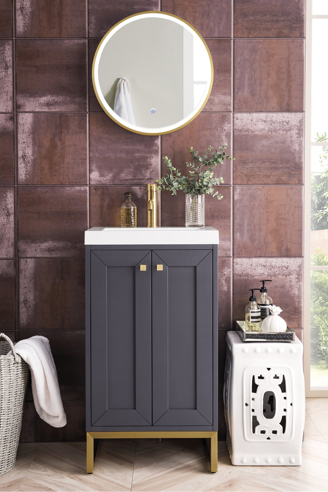 Contemporary solid-wood single-sink vanity in dark gray with a white stone top, brass faucet and gold legs, set against rust-toned textured tile with a round gold mirror and decorative vase and soap dispensers.