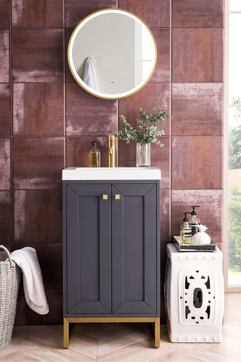 Contemporary solid-wood single-sink vanity in dark gray with a white stone top, brass faucet and gold legs, set against rust-toned textured tile with a round gold mirror and decorative vase and soap dispensers.