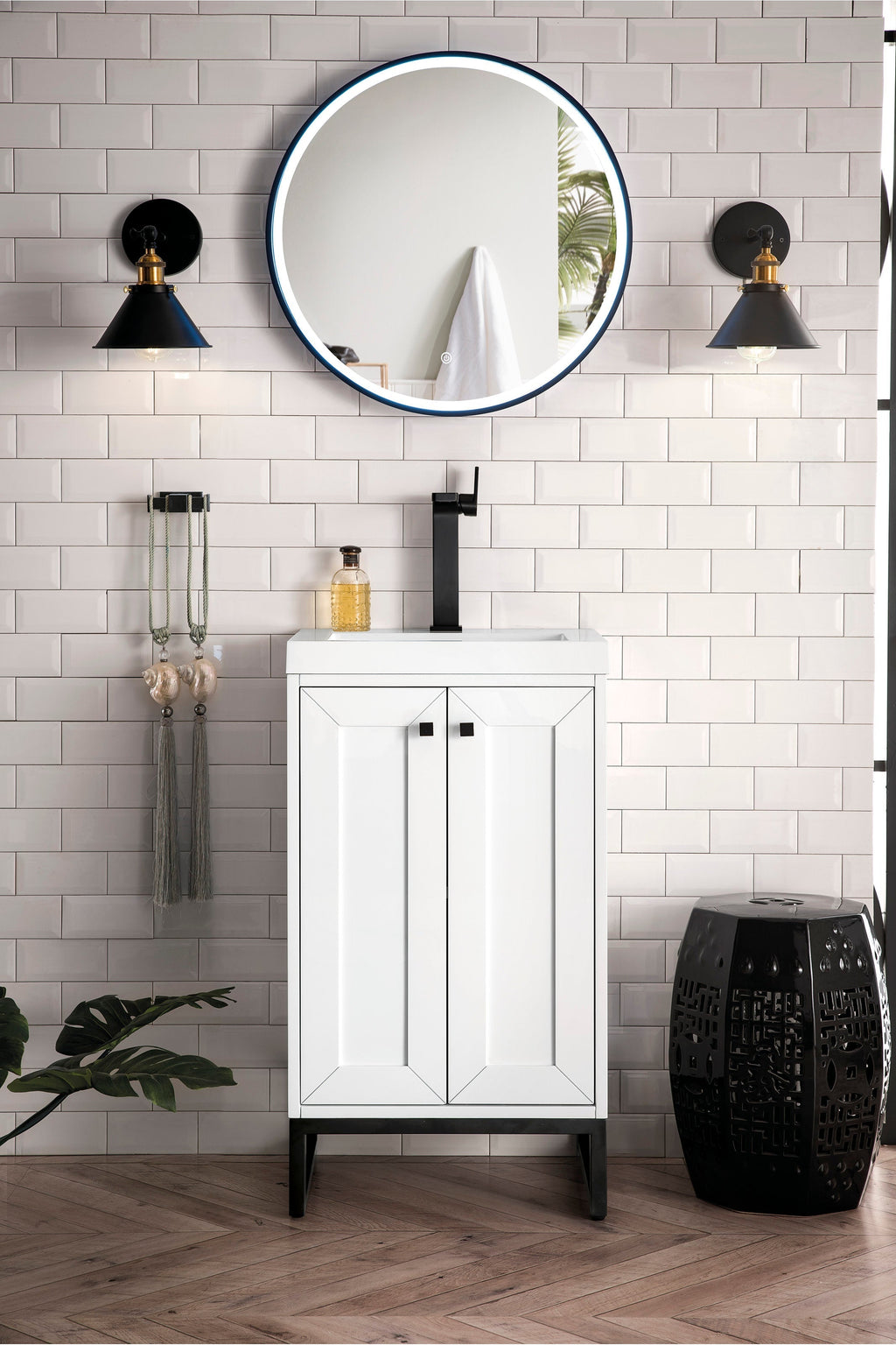 Modern white solid-wood single-sink bathroom vanity with a stone top and matte black faucet, shown with a round illuminated mirror, black wall sconces, subway tile wall and a decorative black stool.
