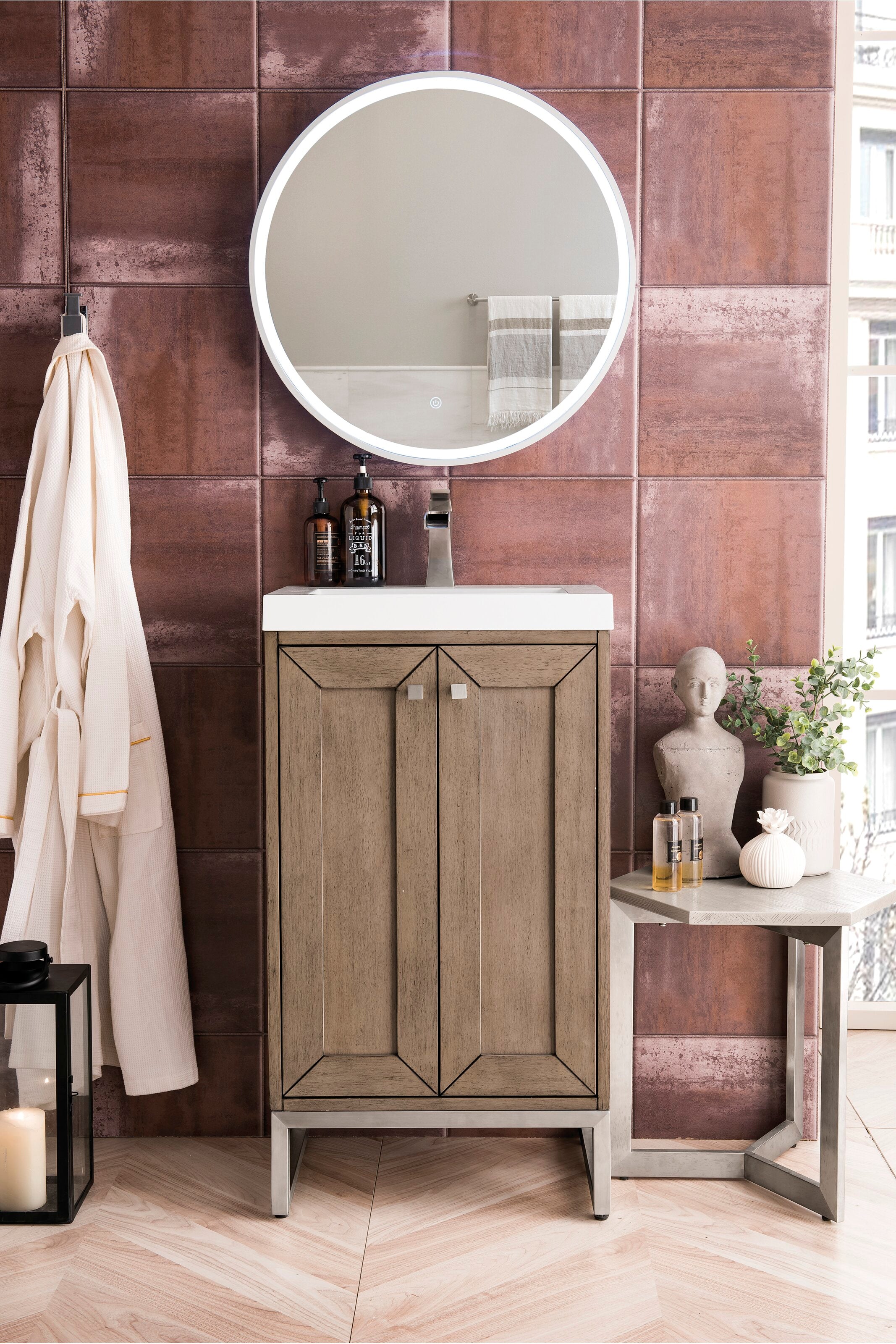 Modern freestanding solid-wood bathroom vanity in a stone finish with a white stone top and integrated sink, chrome faucet and illuminated round mirror, styled with soap dispensers, robe and side-table decor.