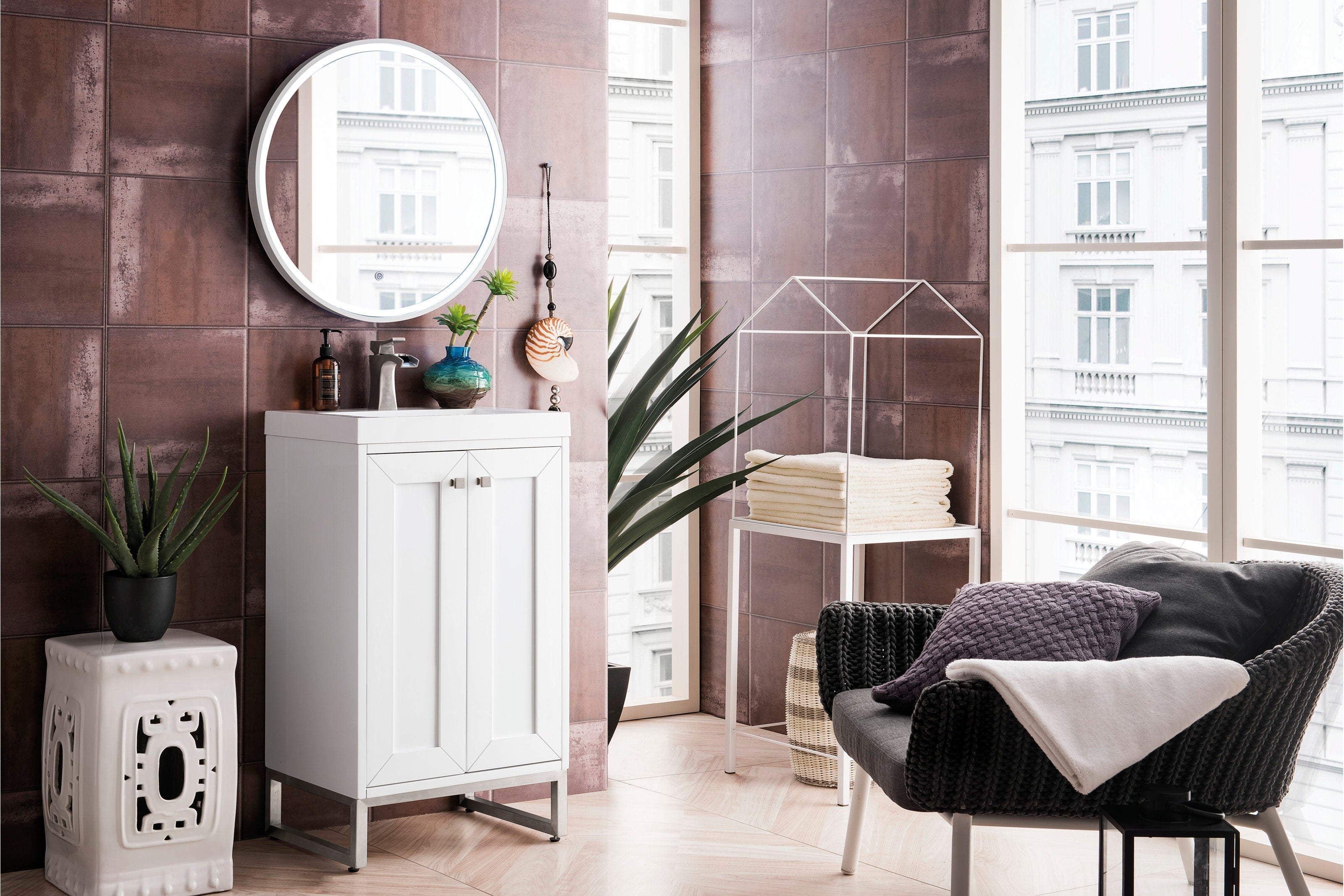 Freestanding white solid-wood bathroom vanity with a stone top and ceramic sink, brushed-nickel faucet and round mirror on a brown tiled wall, styled with potted plants, folded towels and a woven armchair nearby.