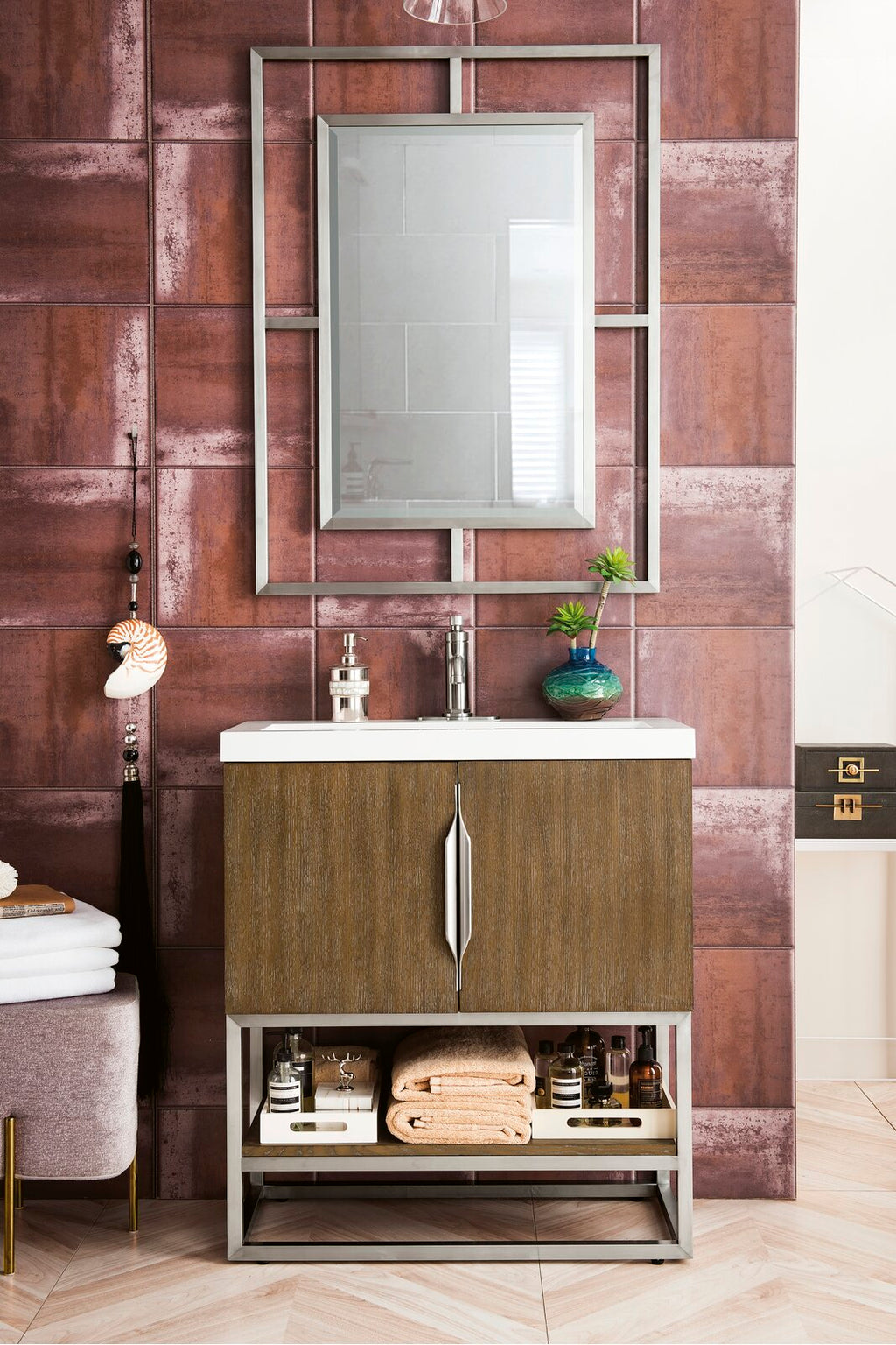 Modern solid wood bathroom vanity with a white stone top, chrome faucet and integrated sink, vertical metal handles, open lower shelf holding folded towels and bottles, framed rectangular mirror and rust-red tiled wall backdrop.