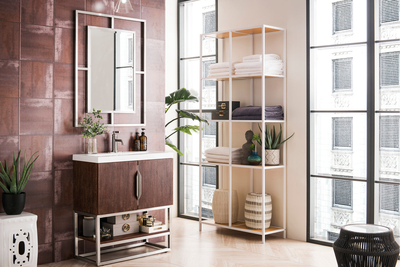 Freestanding modern solid wood single-sink vanity in dark wood with a white stone top, metal base and framed mirror, styled with faucet and bottles; shown next to open shelving with towels, baskets and plants.