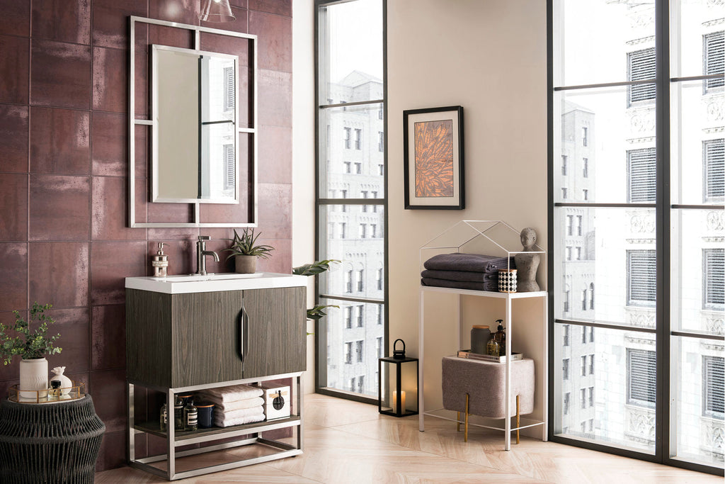 Freestanding modern single-sink bathroom vanity with a white stone top, solid wood stone-finish cabinet on a chrome metal base, framed mirror, burgundy tile wall and styled towels and plants.