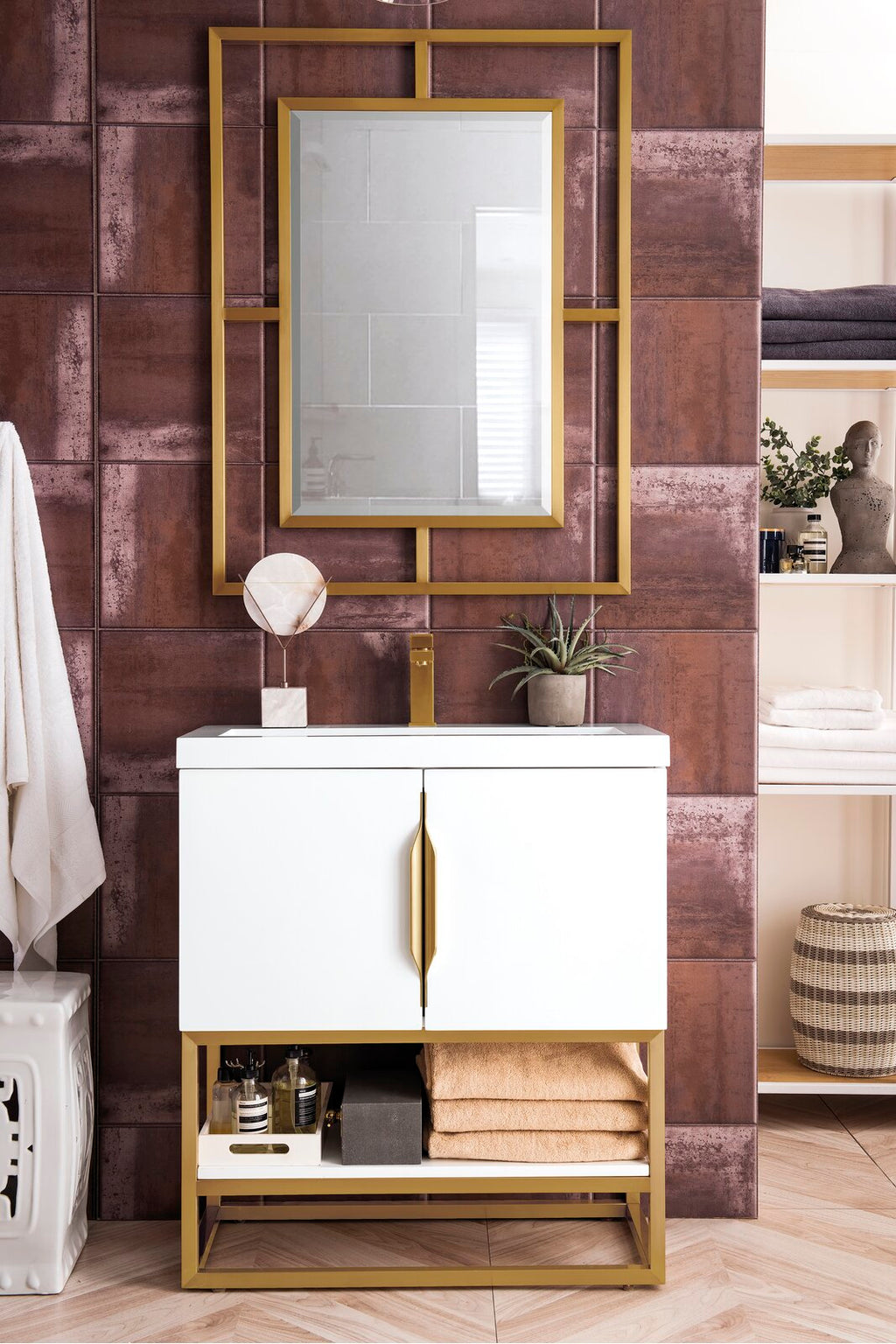 Freestanding modern white single-sink bathroom vanity with a stone countertop, gold faucet and hardware, open lower shelf with folded towels and bottles, and a matching gold-framed mirror against textured brown tile wall.