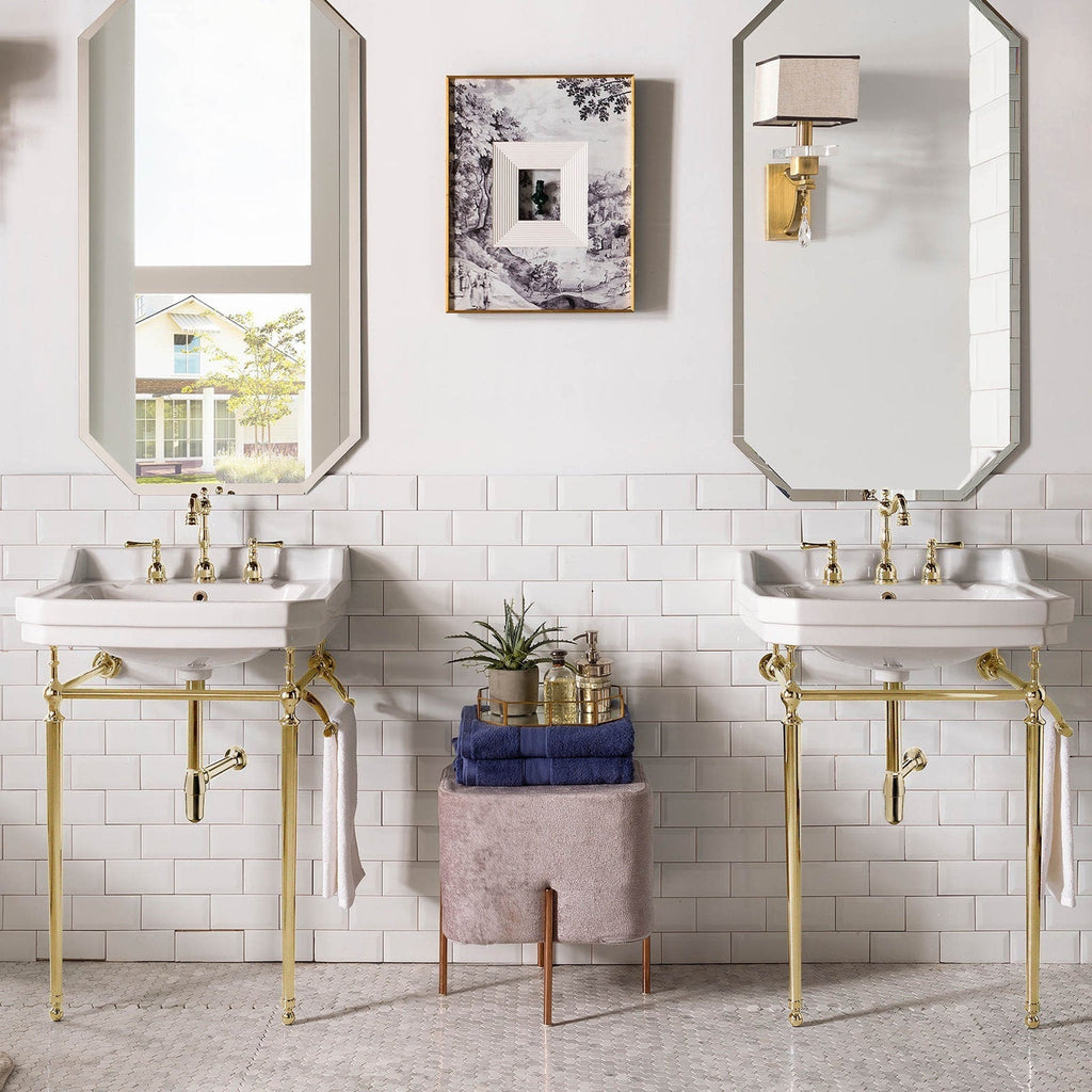 Two white ceramic console sinks with polished brass metal stands and matching brass faucets, mirrored above, flanking a stone-toned upholstered stool stacked with blue towels and bath accessories against white subway tile.