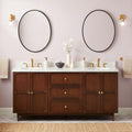 72-inch coastal solid wood double-sink vanity with Carrara marble top, two oval undermount sinks, brass faucets and knobs, round black mirrors, brass wall sconces and decorative vases against a soft pink wall.