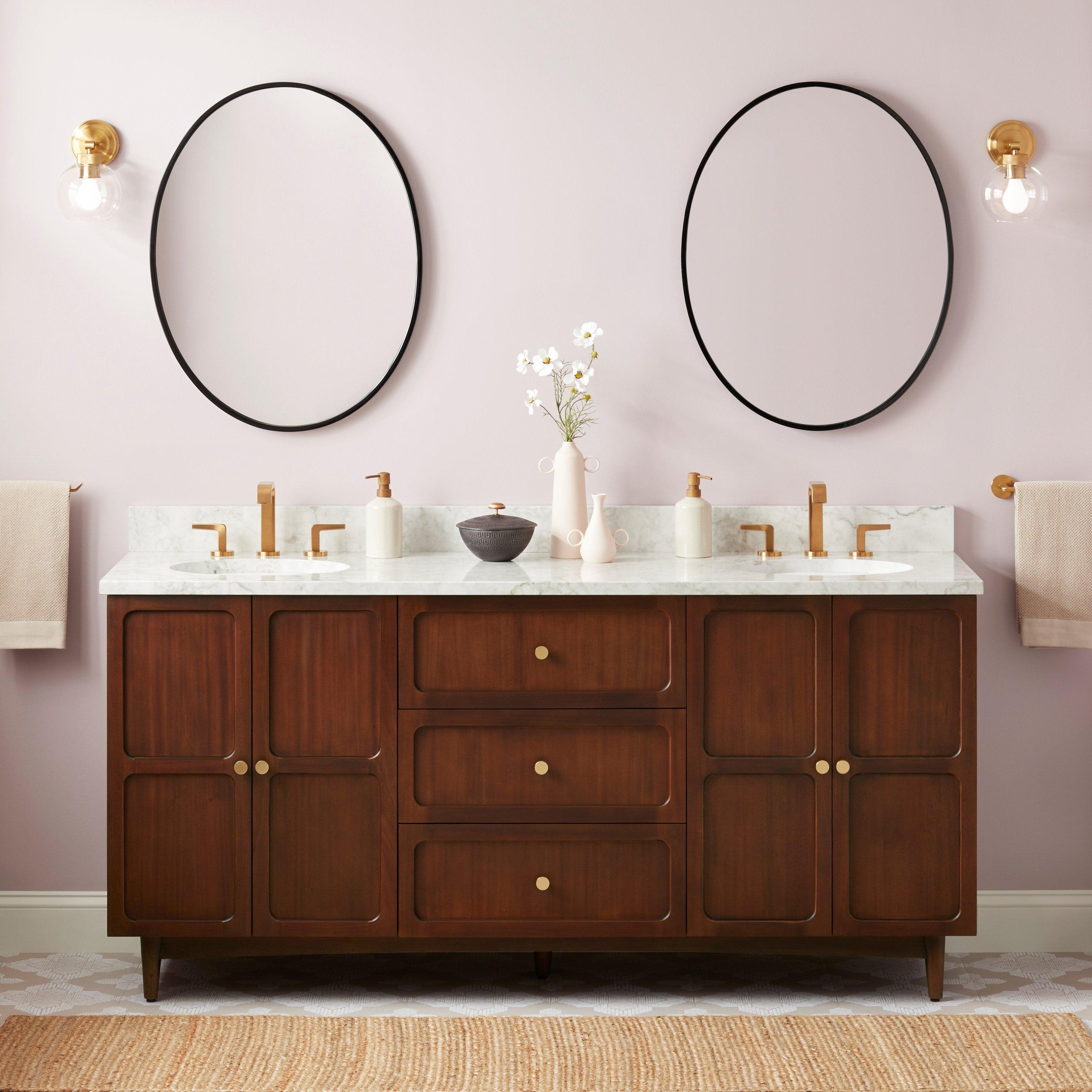 72-inch coastal solid wood double-sink vanity with Carrara marble top, two oval undermount sinks, brass faucets and knobs, round black mirrors, brass wall sconces and decorative vases against a soft pink wall.