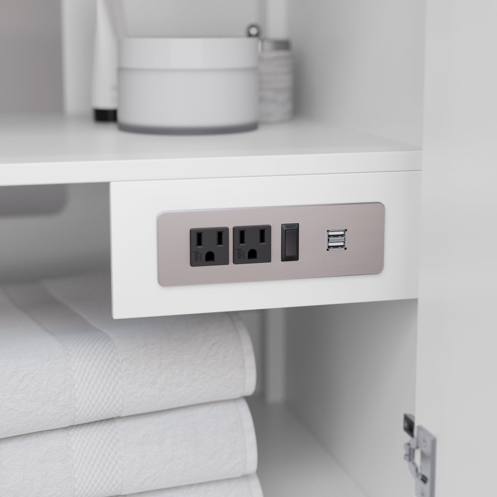 Close-up of a white wooden vanity interior showing a brushed metal power plate with two black electrical outlets, a reset switch and dual USB ports, with folded white towels below and toiletries on the upper shelf.