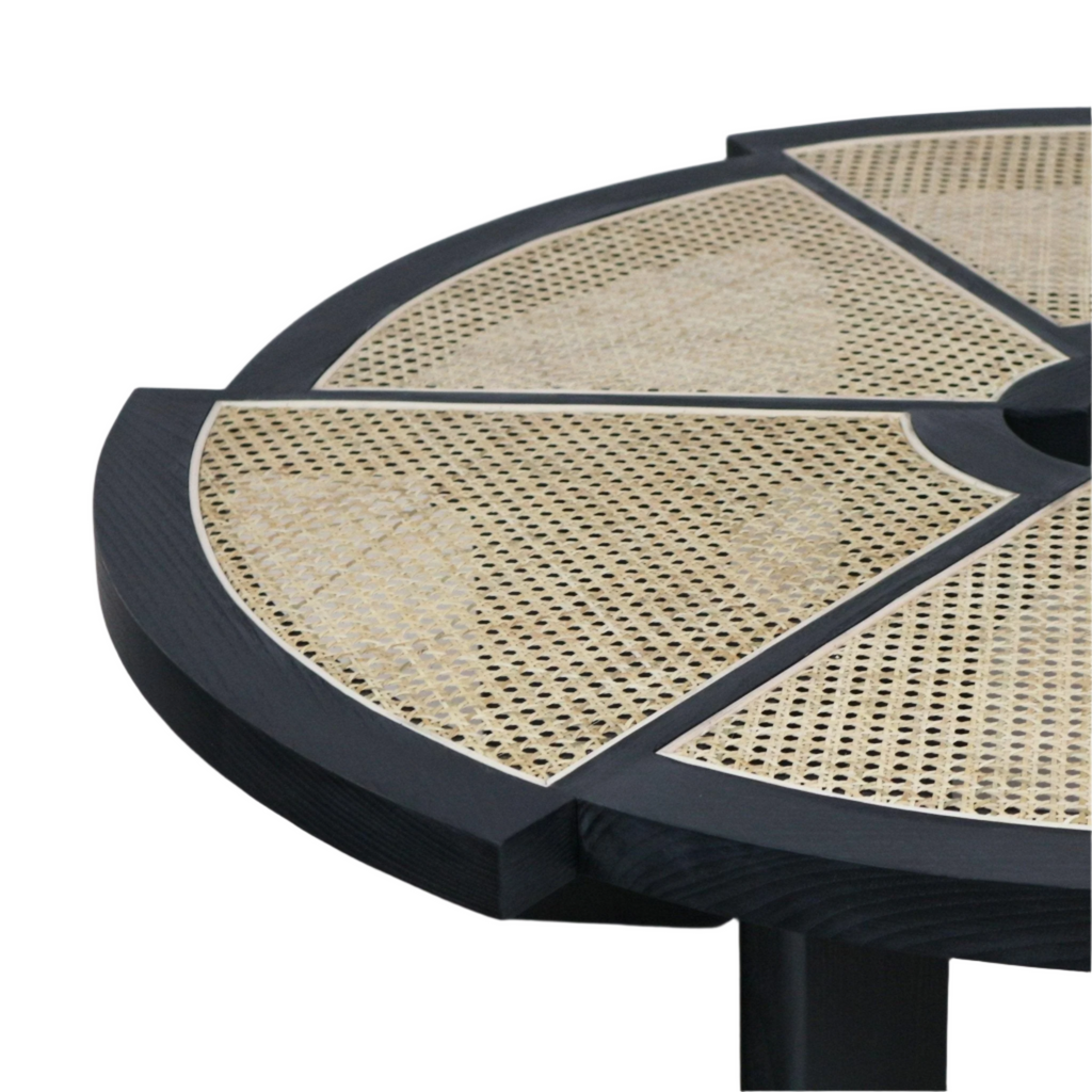 Close-up of a round modern coffee table top showing a black stained wood frame with natural woven cane panels arranged in segmented radial sections and a portion of the black pedestal leg.