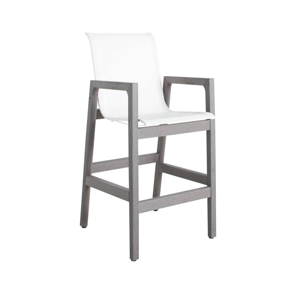 Coastal teak outdoor bar stool with a weathered grey teak frame, white sling seat and integrated armrests, counter-height design with square legs and built-in footrest.