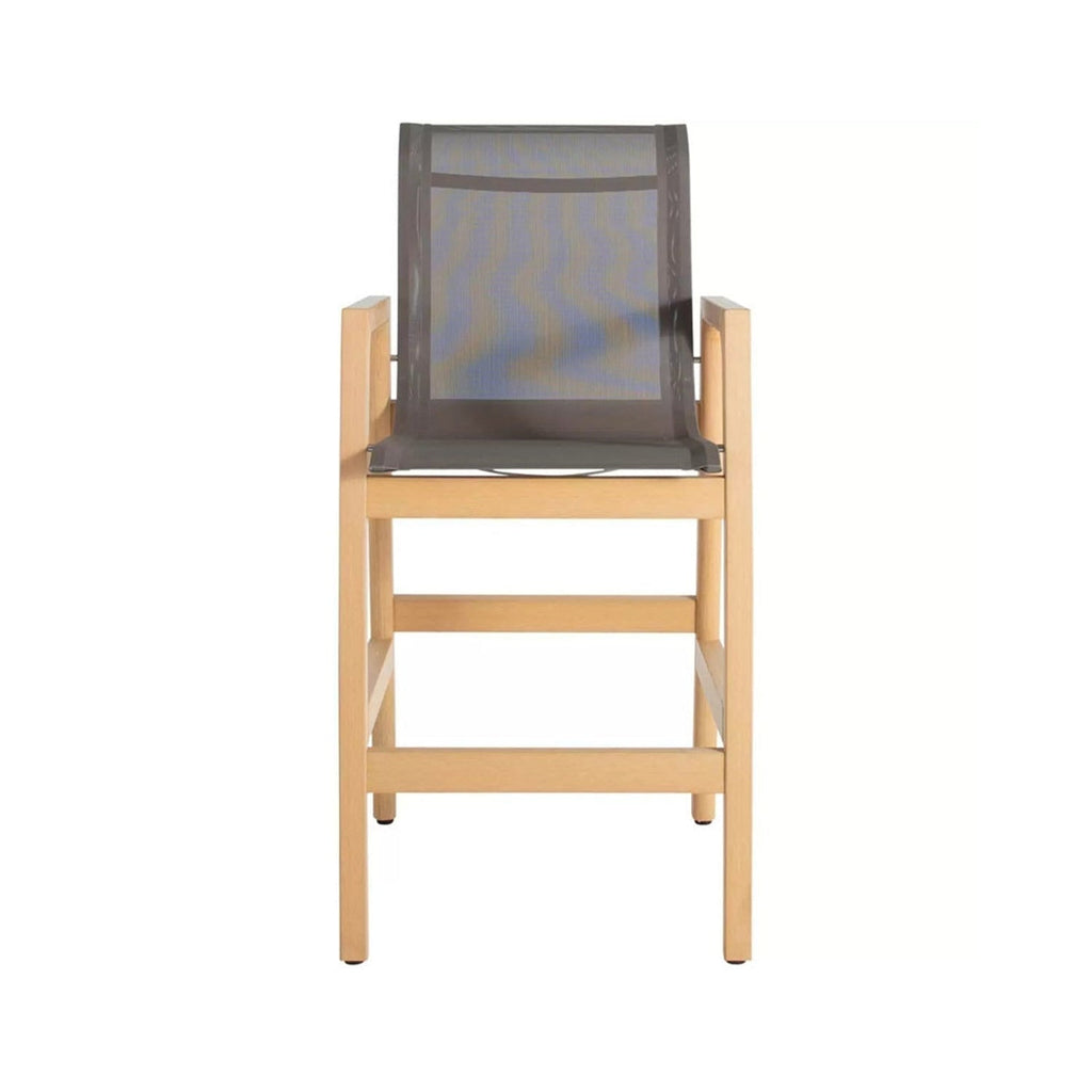 Front view of a teak outdoor bar stool with a natural finish wooden frame, gray mesh sling seat and back, squared armrests and integrated footrest, coastal-style counter-height seating.