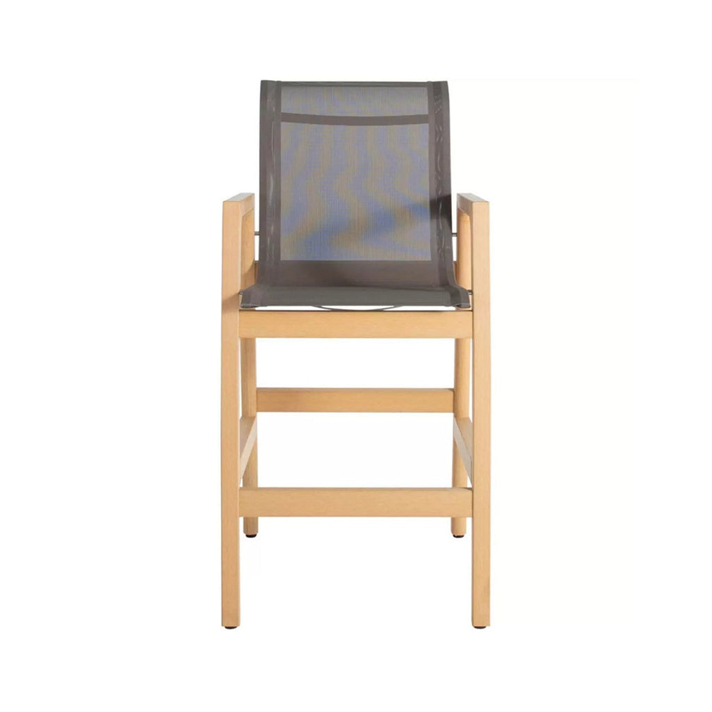 Front view of a teak outdoor bar stool with a natural finish wooden frame, gray mesh sling seat and back, squared armrests and integrated footrest, coastal-style counter-height seating.