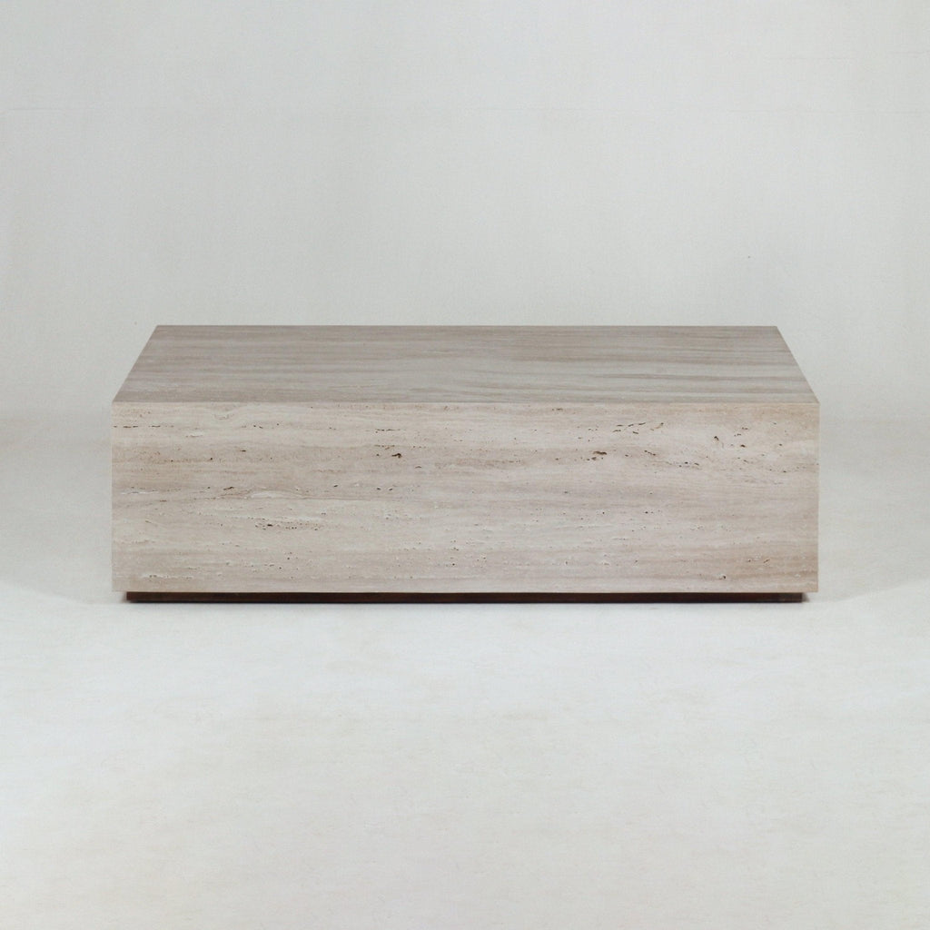 Solid rectangular travertine coffee table in natural beige with subtle horizontal veining, sitting low on a slim dark plinth against a neutral studio background.