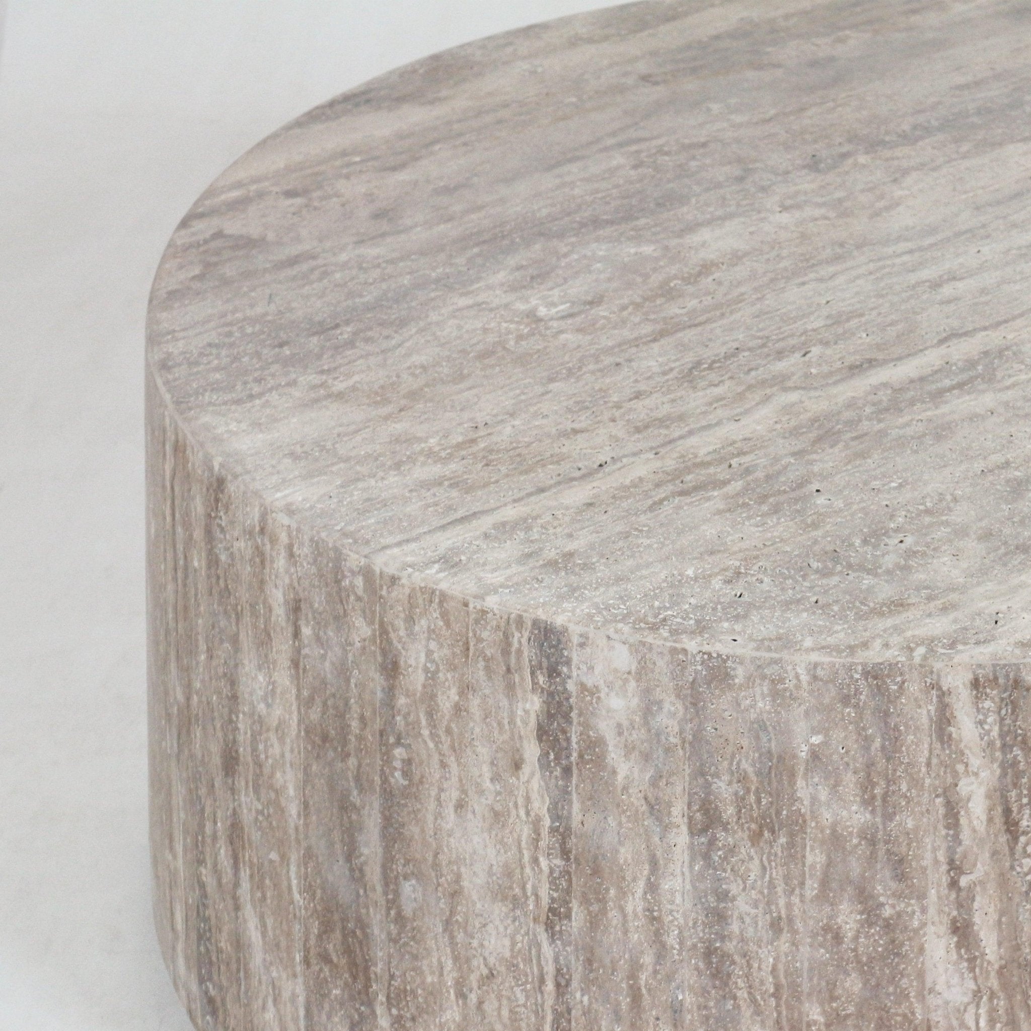 Close-up of a round travertine coffee table showing the natural beige stone top and textured vertical striations on the side.