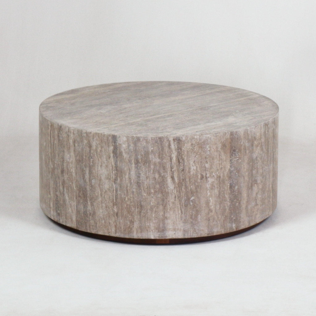 Low round travertine coffee table in natural beige with a solid cylindrical form, smooth stone top and a subtle dark plinth base, modern minimalist style for living or outdoor spaces.