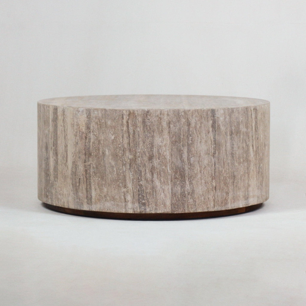 Round travertine coffee table in natural beige with vertical stone veining, low cylindrical drum form, and a slim recessed dark wood plinth, shown on a neutral background.