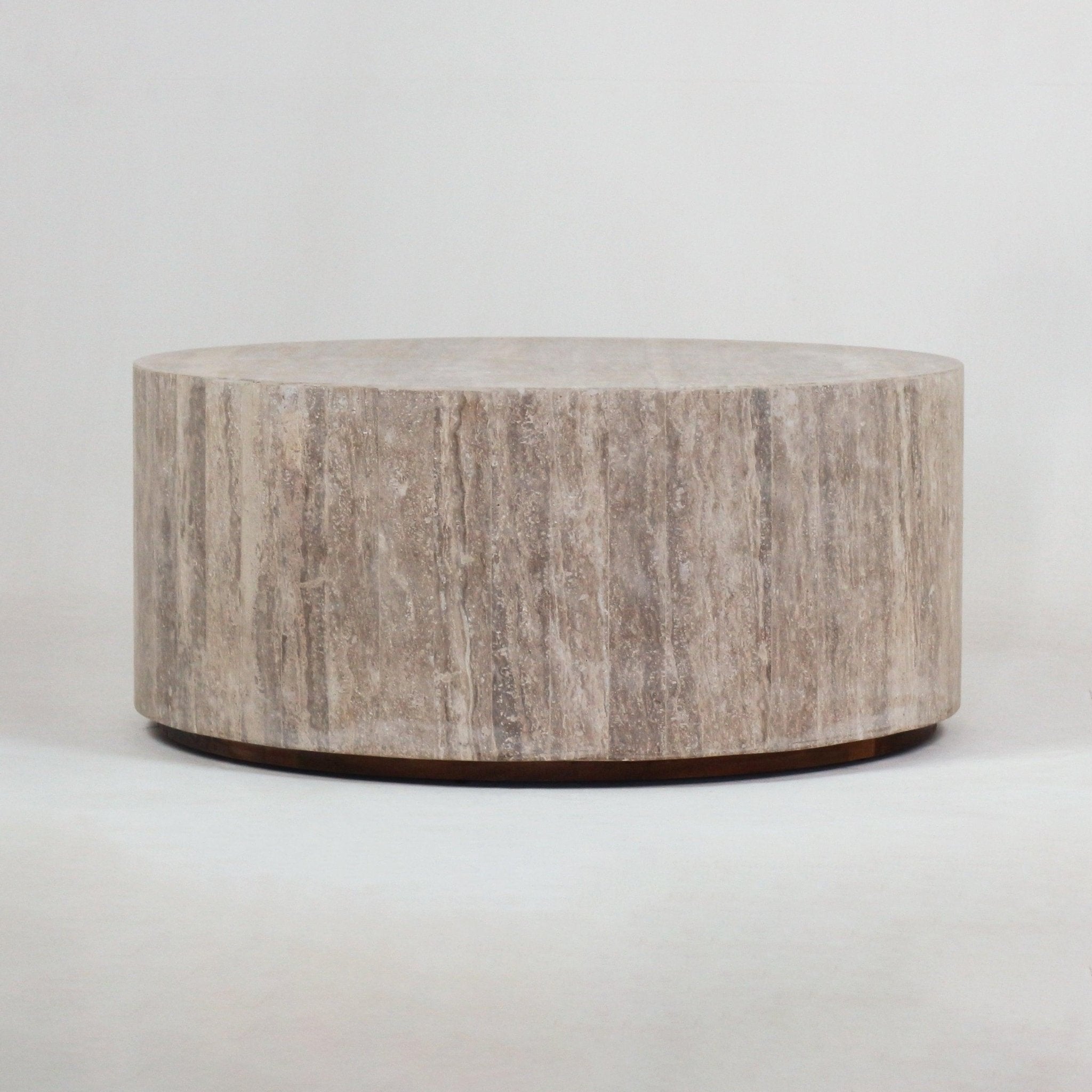 Round travertine coffee table in natural beige with vertical stone veining, low cylindrical drum form, and a slim recessed dark wood plinth, shown on a neutral background.