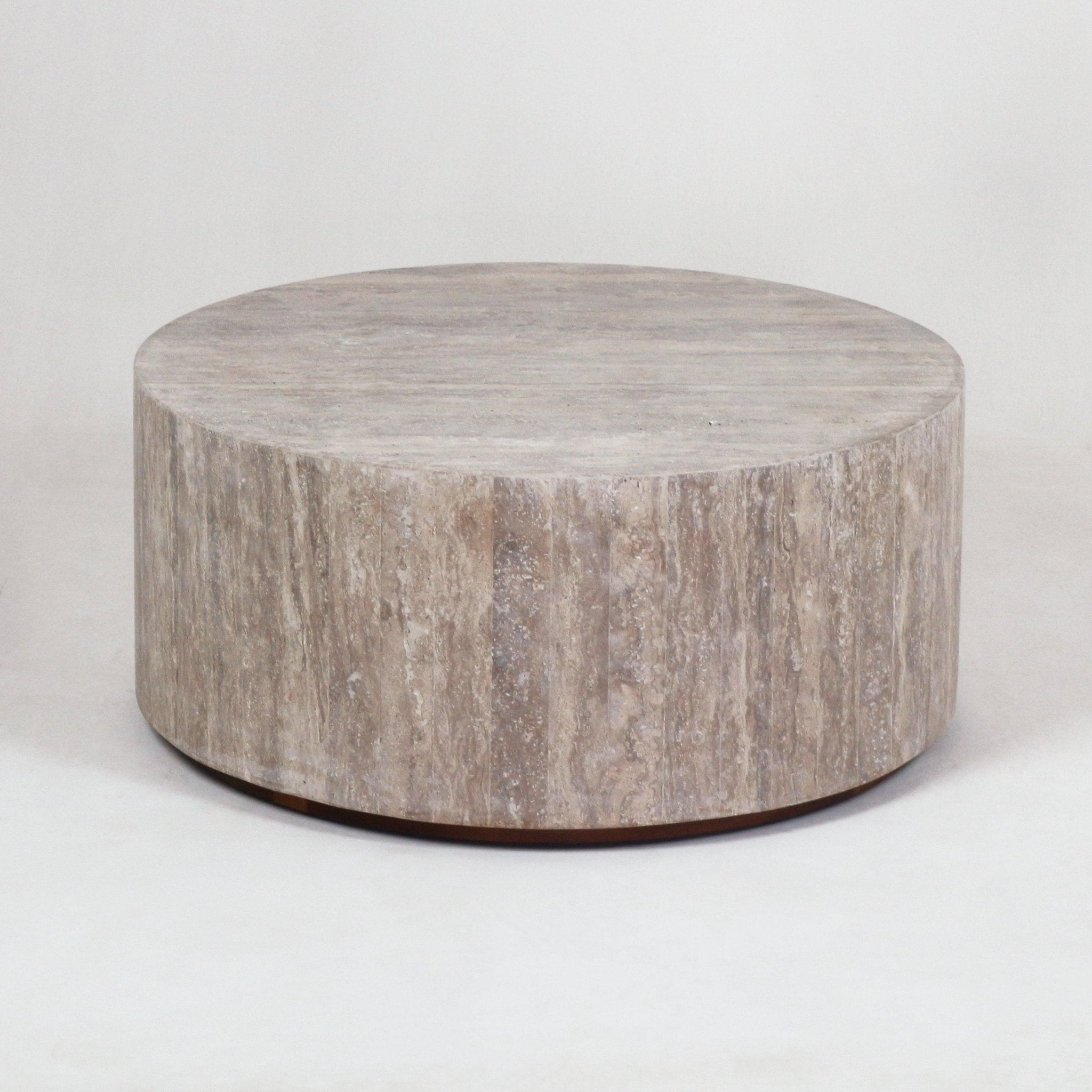 Round, low-profile travertine coffee table in natural beige stone with a cylindrical drum shape, subtle vertical veining and a slightly recessed dark base.