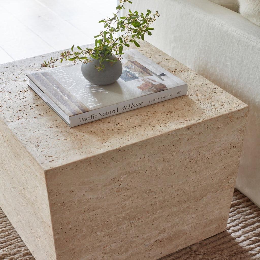 Natural beige travertine cube nightstand with visible stone texture, styled with a coffee-table book and a small gray vase holding green sprigs next to a cream sofa and textured rug.