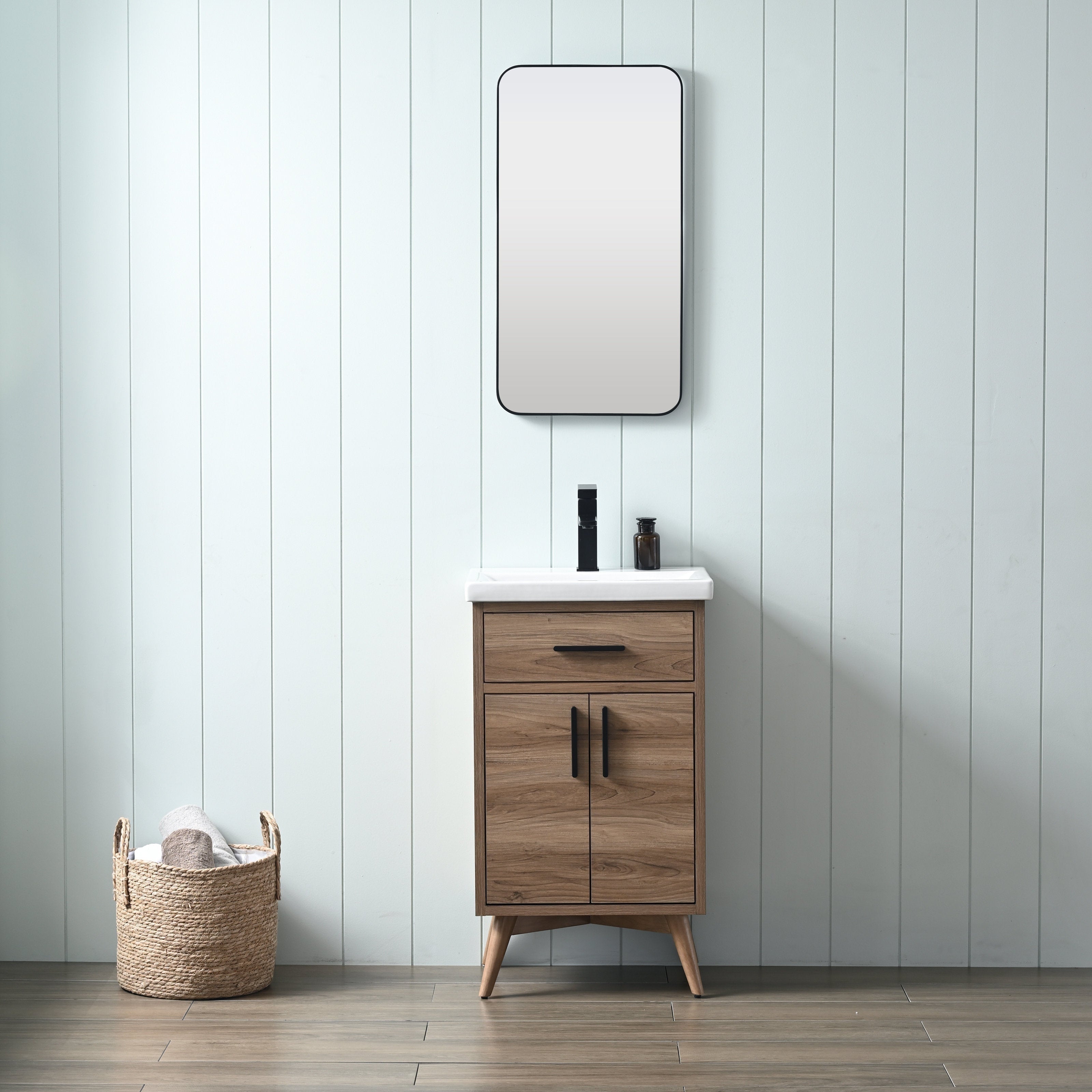Freestanding modern solid wood bathroom vanity in natural walnut with a white ceramic sink, black faucet and matching black handles, rounded rectangular mirror above and woven towel basket against a pale shiplap wall.