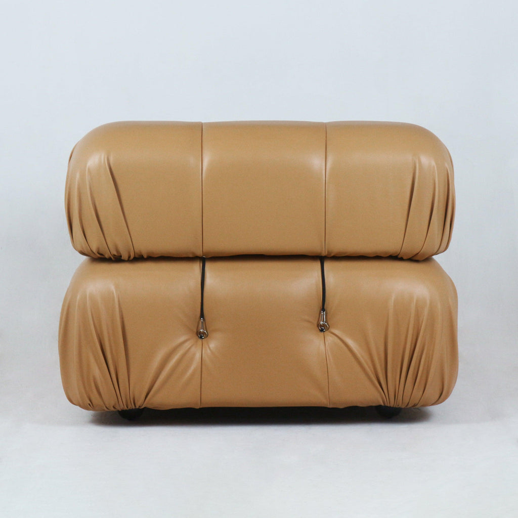 Front view of a tan leather modern sofa with two stacked rounded cushions, gathered corner pleats, tufted buttons with metal strap hardware and a low profile on short feet.