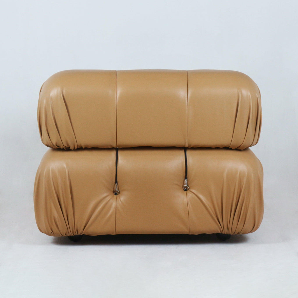 Front view of a tan leather modern sofa with two stacked rounded cushions, gathered corner pleats, tufted buttons with metal strap hardware and a low profile on short feet.