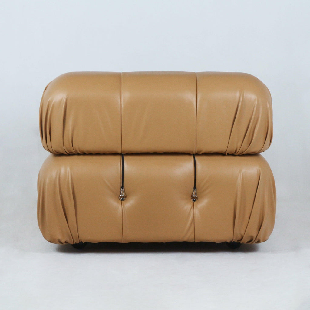 Front view of a modern tan leather sofa with two stacked rounded cushions, subtle tufting and black strap hardware on a white backdrop.