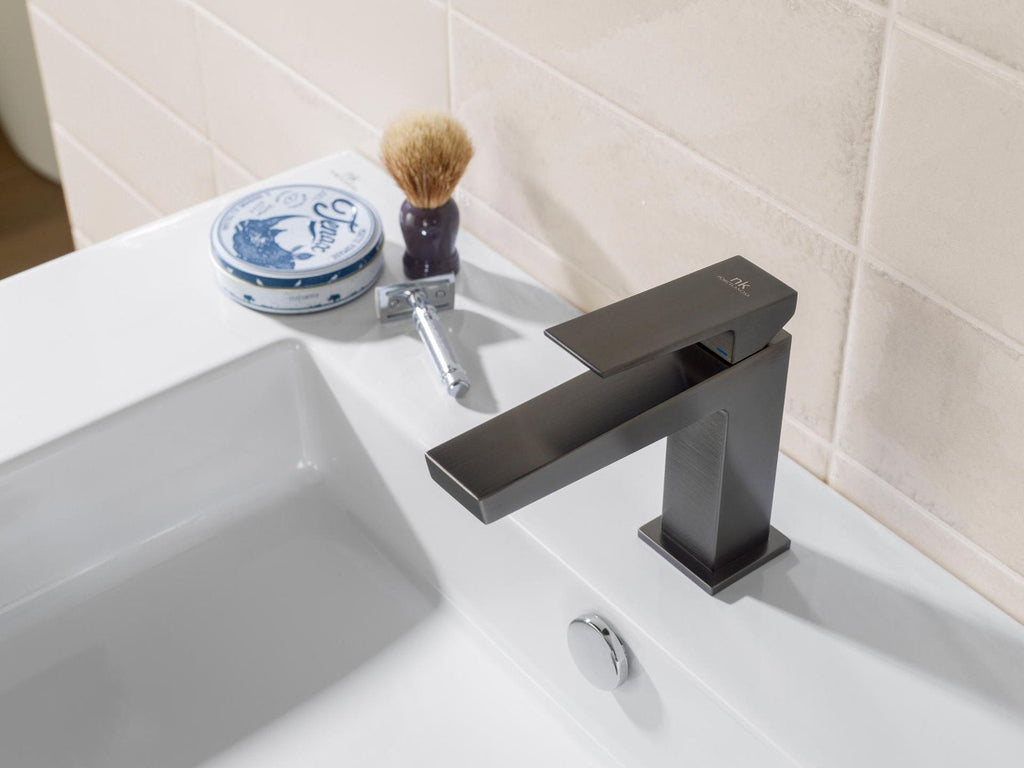 Close-up of a modern matte dark single-handle faucet on a stone-colored vanity top above a white rectangular sink, with a chrome drain stopper and nearby shaving brush, razor and blue round soap tin against beige tile.