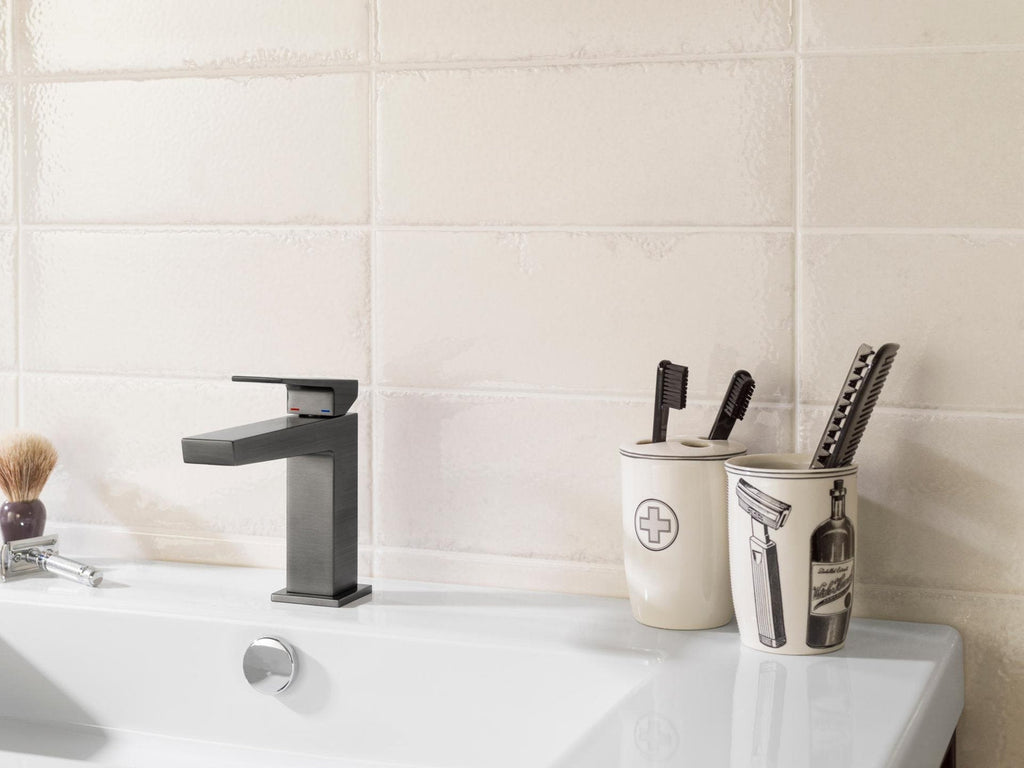 Close-up of a stone-colored vanity top with a modern square dark chrome faucet beside a white rectangular sink, two ceramic toothbrush holders with black brushes, and a shaving brush and razor on the left.