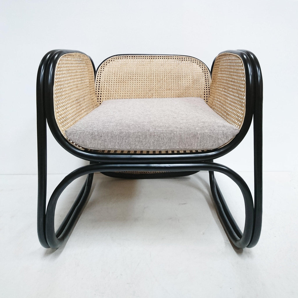 Front view of a modern lounge chair with a black curved metal frame, natural woven cane back and sides, and a light gray fabric seat cushion.