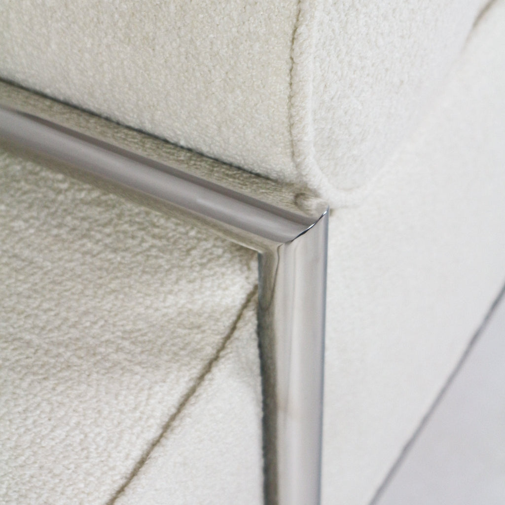 Close-up of a cream boucle upholstered end-of-bed bench corner showing textured boucle fabric and a polished stainless steel frame with a mitered metal joint.