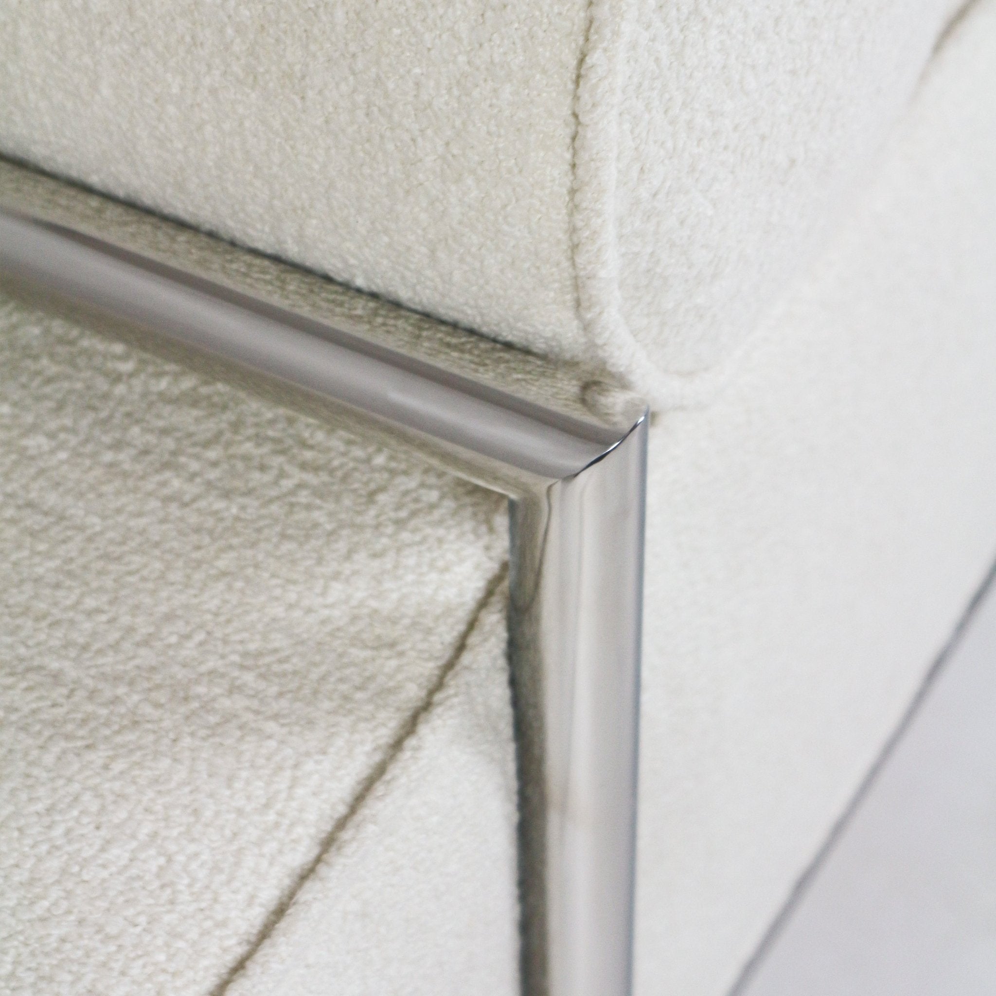 Close-up of a cream boucle upholstered end-of-bed bench corner showing textured boucle fabric and a polished stainless steel frame with a mitered metal joint.