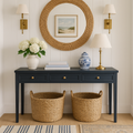 Navy solid wood console table with three drawers and brass knobs, styled beneath a round woven rattan mirror with brass sconces, topped with a lamp, blue-and-white ceramic jar and white hydrangeas, with two woven storage baskets below.