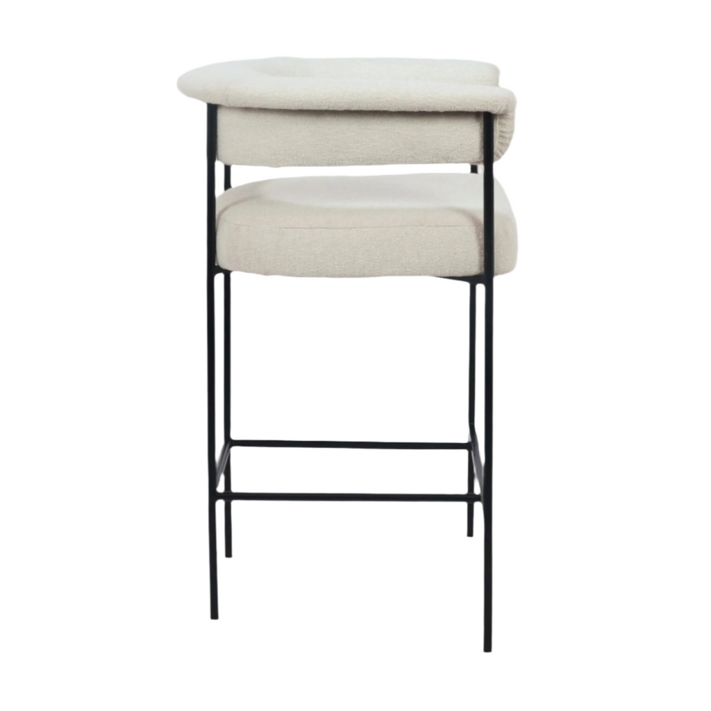 Cream boucle upholstered counter stool with a low rounded back and padded seat, supported by a slim matte black metal frame with integrated footrest, shown on a white background in a modern minimalist style.