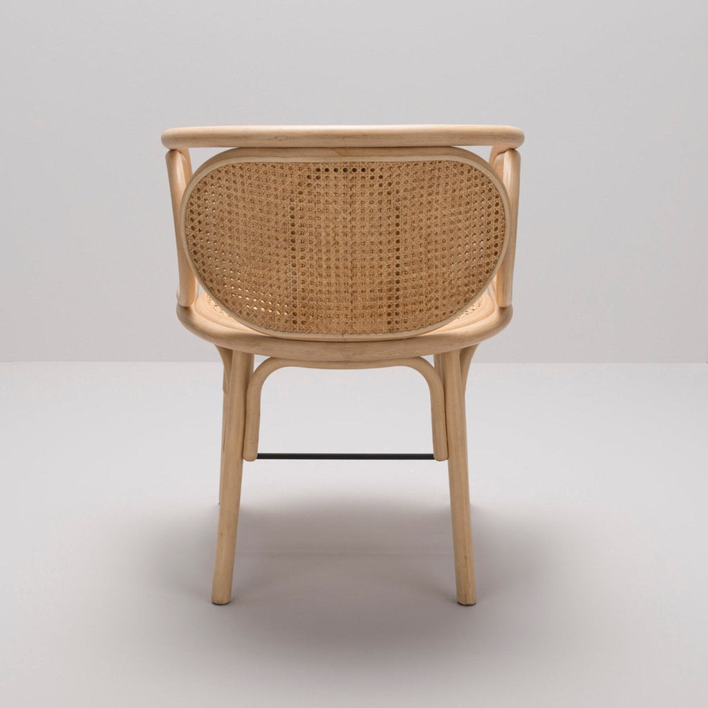 Rear view of a natural wood rattan dining chair with an oval cane-woven back, curved bentwood frame and a black metal stretcher bar, shown in a boho-inspired coastal style.