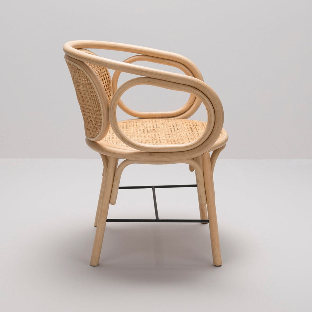 Side view of a natural wood rattan dining chair with looped curved arms, cane-woven seat and back, tapered legs and a black metal footrest, photographed against a light neutral background.