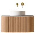 Floating fluted teak wood bathroom vanity with a light stone top and round white ceramic vessel sink, paired with a wall-mounted brushed brass spout and round single-lever control.