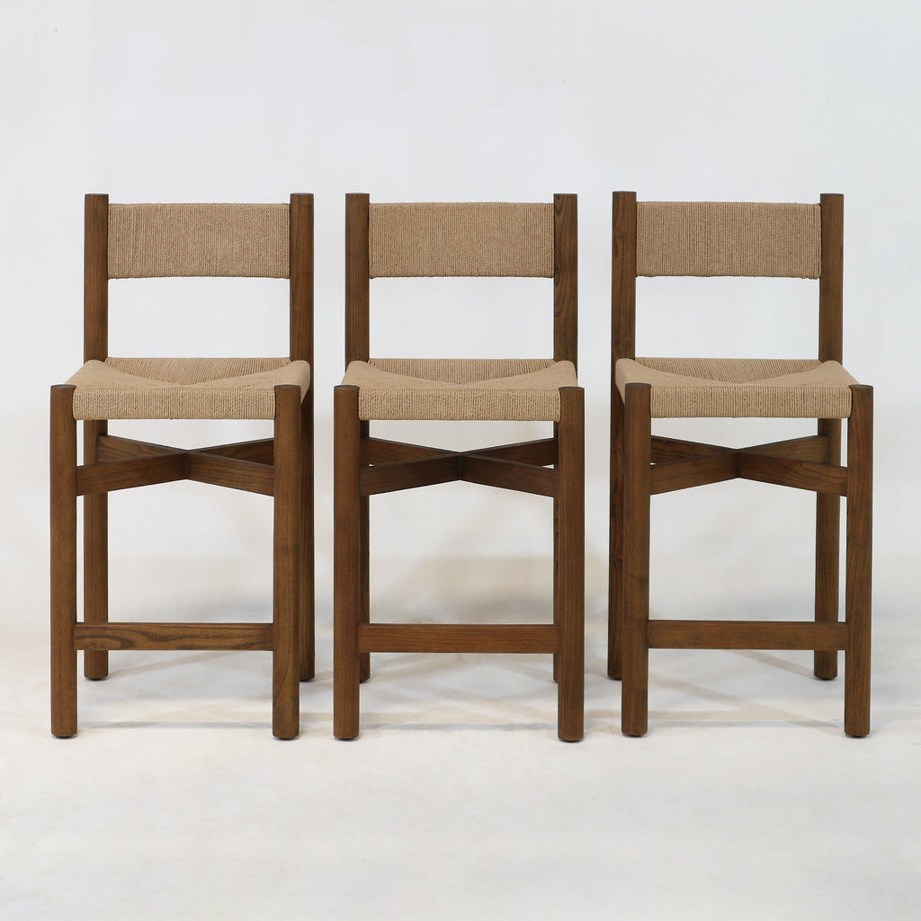 Three walnut-finished solid wood counter-height stools with natural papercord-woven seats and backs, square legs and X-shaped stretchers in a modern coastal style, photographed against a plain white background.