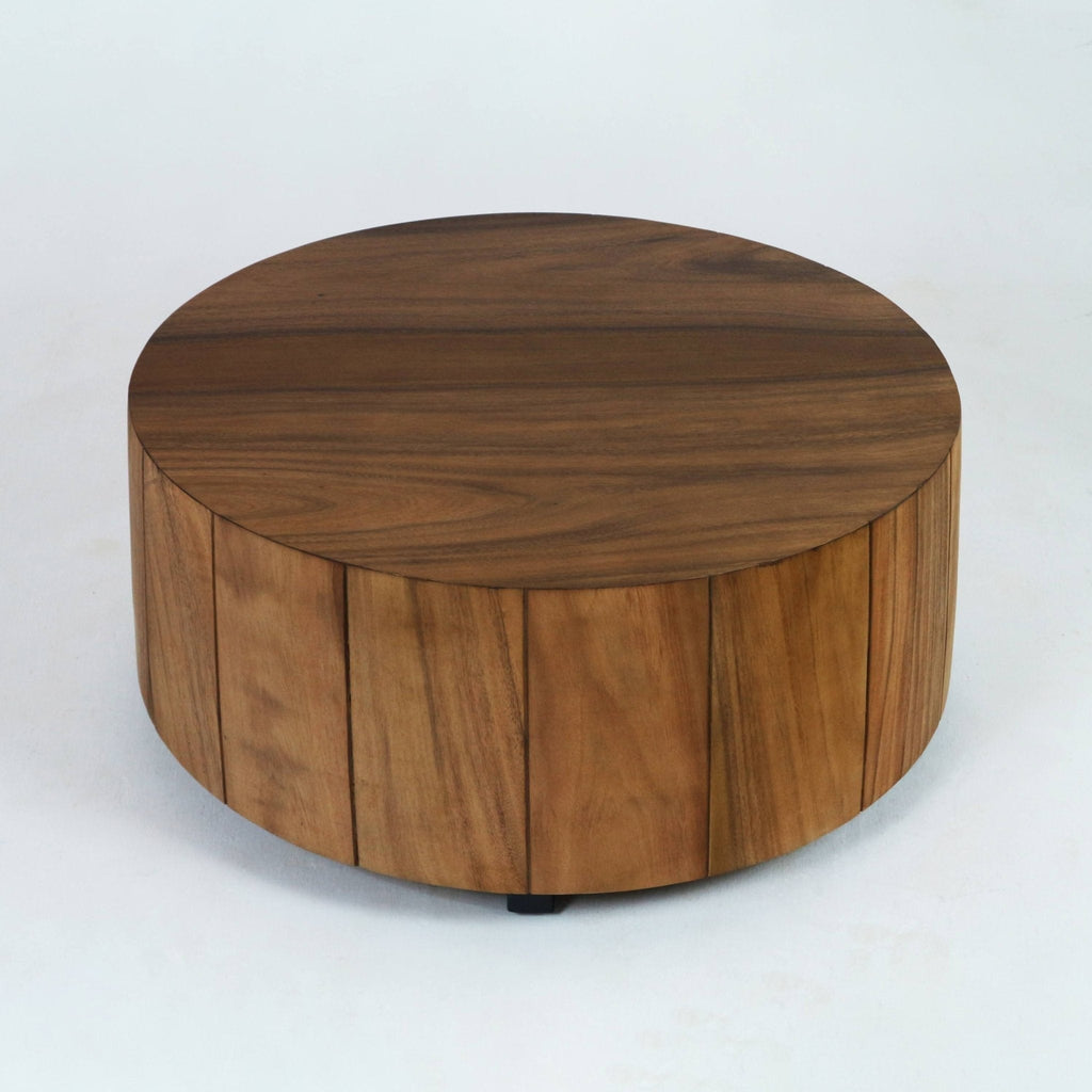 Low, round solid wood coffee table with a warm natural wood finish, cylindrical body of vertical planks and a smooth top, shown resting on small black feet against a white studio background