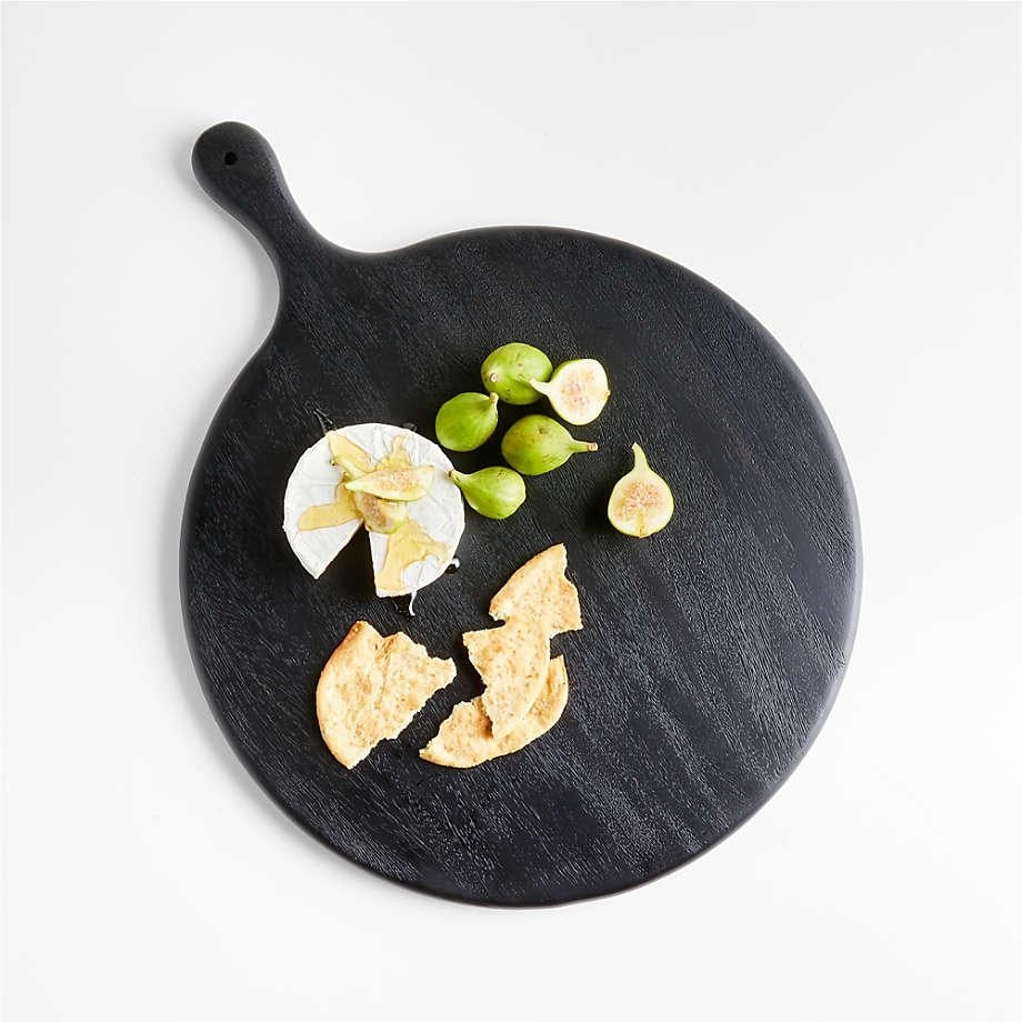 Round black oak serving board with handle topped with halved green figs, a wedge of brie drizzled with honey and broken crackers on a white background.