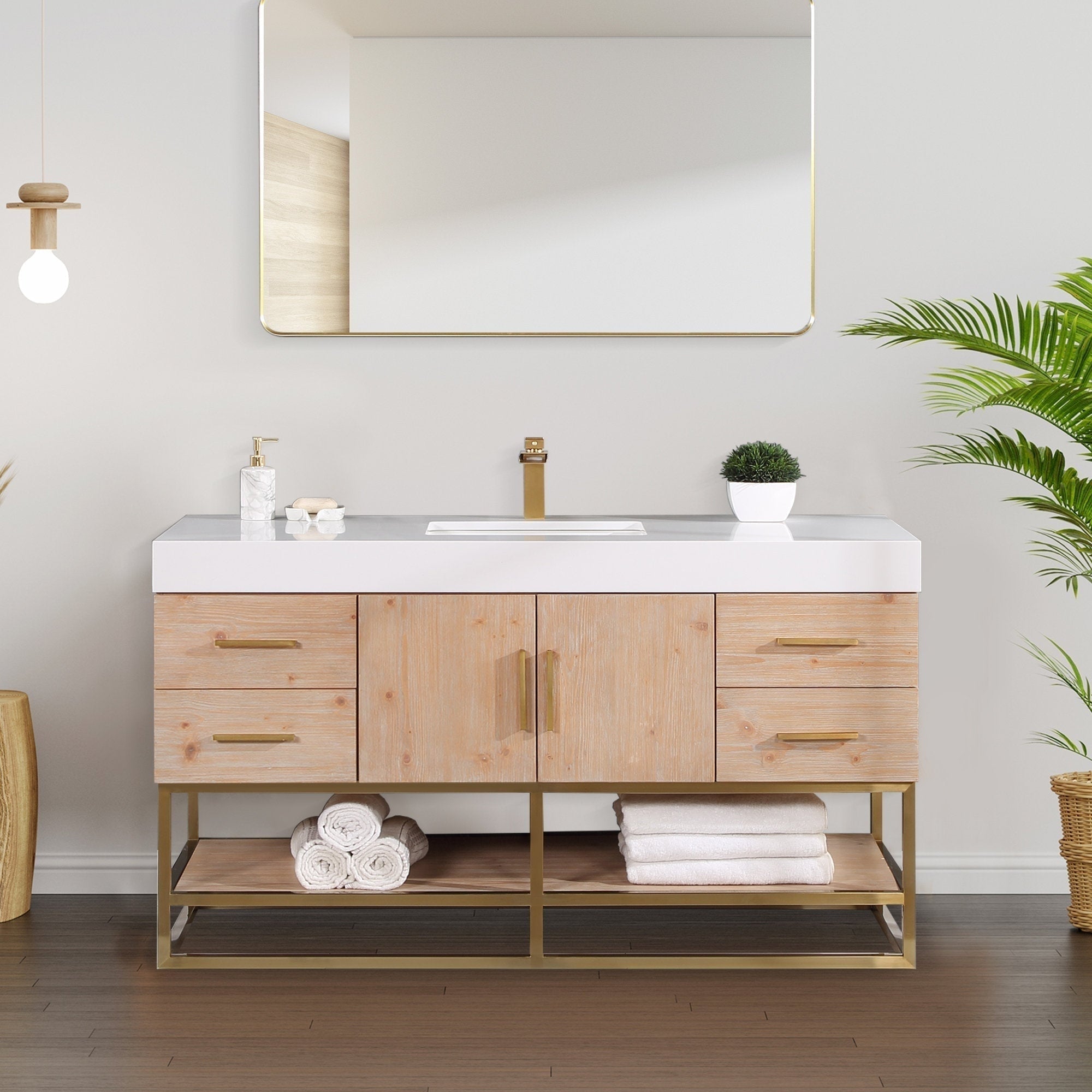 Modern 60-inch light natural wood bathroom vanity with white composite stone top, rectangular undermount sink, brass-finish faucet and handles, open lower shelf with rolled towels, gold-framed mirror and potted plant.