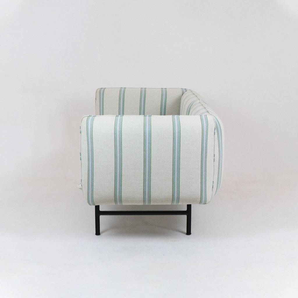 Side view of an outdoor sectional sofa upholstered in light neutral fabric with vertical green-blue stripes, featuring low black metal legs and rounded arm and back against a plain white background.